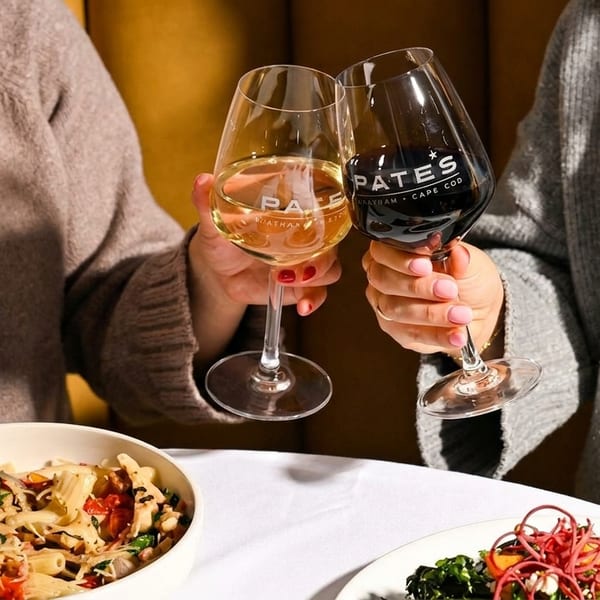 Two people clinking wine glasses with the Pates logo, alongside bowls of pasta and salad.