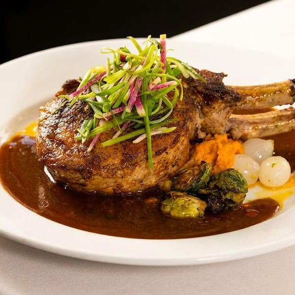 A large bone-in pork chop with gravy, brussels sprouts, pearl onions, and sweet potato mash.