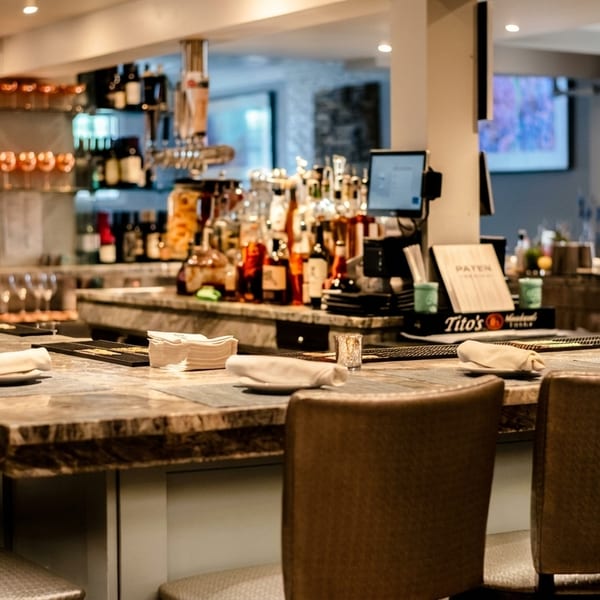 Empty bar area with stools, set plates, and bottles of liquor and wine on shelves.