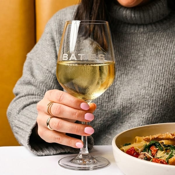 A person in a gray sweater holds a Bates wine glass next to a bowl of pasta.
