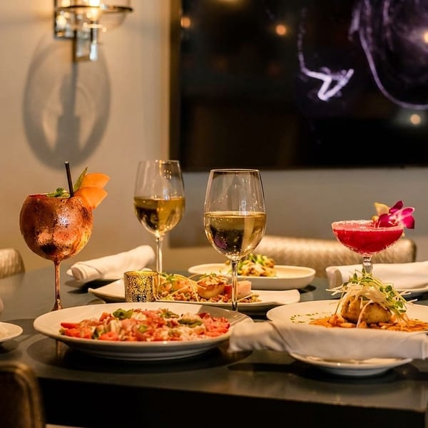 A table set with multiple plates of food, cocktails, and wine glasses in a dimly lit restaurant.