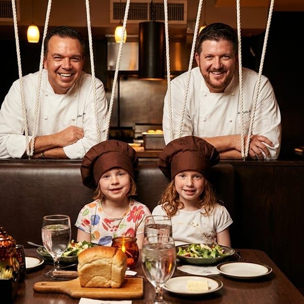 Two smiling chefs behind a booth with two young girls in chef hats at a restaurant table.