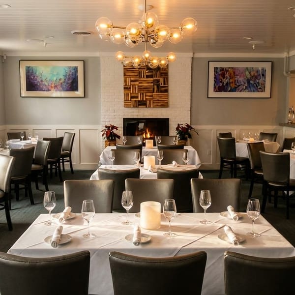 Elegant dining room with white-clothed tables, dark chairs, a lit fireplace, and a bar area.