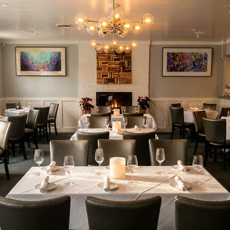 Elegant dining room with white-clothed tables, dark chairs, a lit fireplace, and a bar area.