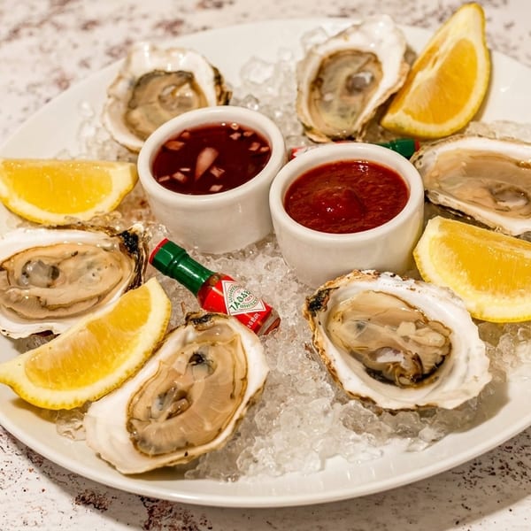 A plate of fresh oysters on ice with lemon wedges, cocktail sauce, mignonette, and Tabasco.