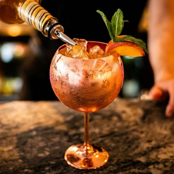 Bartender pouring a drink into an ornate copper goblet with ice, mint, and a peach slice.